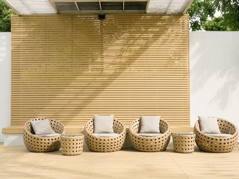 Outdoor seating area with five round wicker chairs and two small wicker tables on a wooden deck, set against a slatted wooden privacy wall.