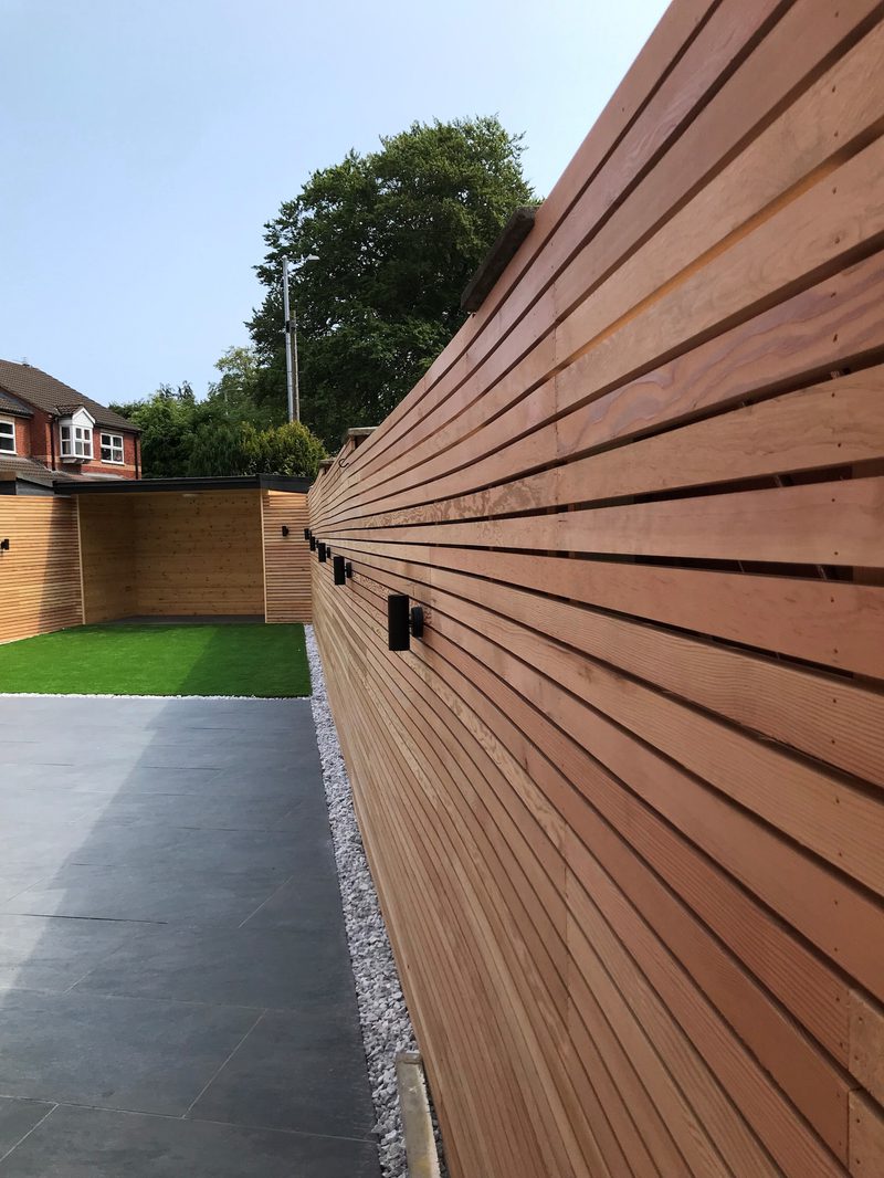 Long wooden slat fence along a paved area with a gravel border; black wall lights attached. Behind it, a small wooden shed, green lawn and trees.