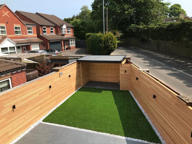 Enclosed backyard with an artificial grass patch bordered by white gravel and wooden fencing; gray patio in front, street and houses beyond.