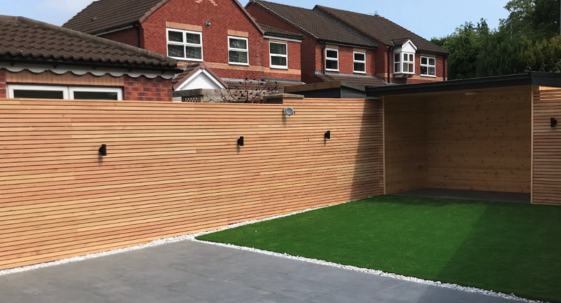 Backyard with horizontal wooden fence, a paved gray patio, a strip of artificial grass, white gravel border, and a covered wooden shed; red brick houses in the background.