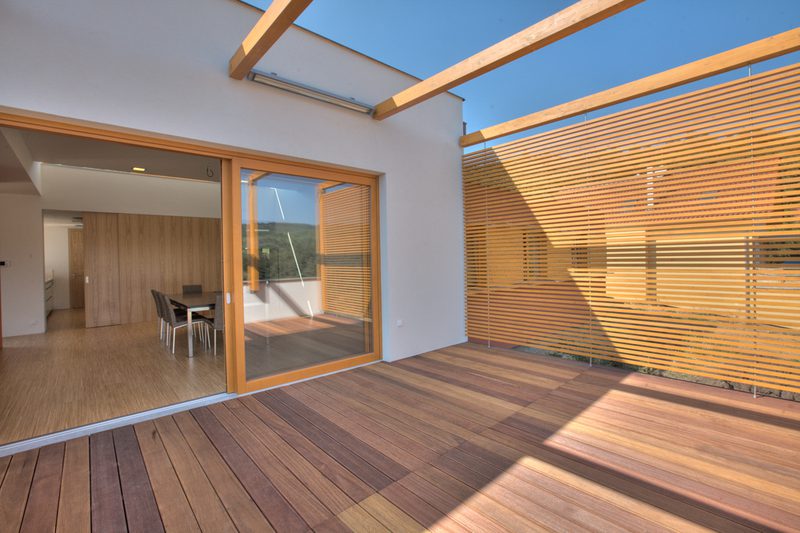 Sunlit wooden deck with a sliding glass door to an interior dining area; a wooden slat screen casts striped shadows on the deck.