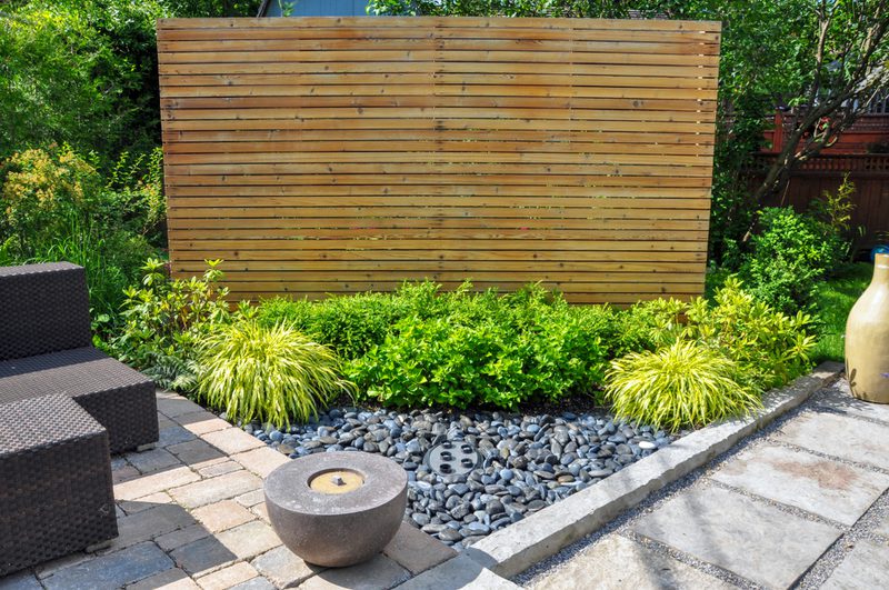 Backyard patio with a tall wooden privacy screen, lush green shrubs, dark river rocks, and wicker seating on pavers beside a large ceramic vase.