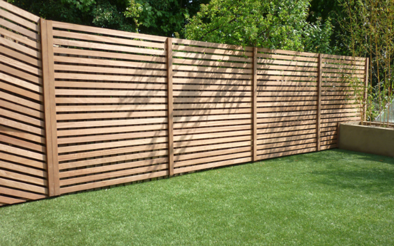 Wooden horizontal slat fence along a green lawn, with tree shadows across the fence and a concrete planter on the right.