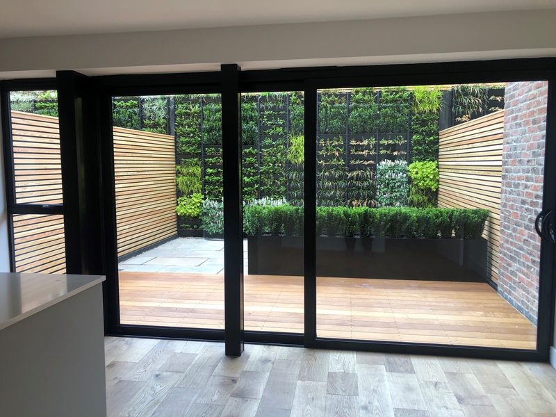 Interior room with large black-framed sliding glass doors opening onto a wooden deck, wooden privacy screens, and a courtyard garden with a vertical plant wall, hedges, and brick.