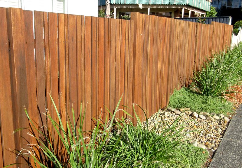 Brown wooden privacy fence with vertical boards, a pebble bed and tall grasses along a sidewalk, with a house and green awning in the background.
