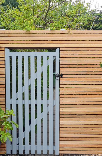 Wooden horizontal slat fence with a gray central gate; dense hedge to the left and a rounded potted tree in a rock-filled pot to the right.