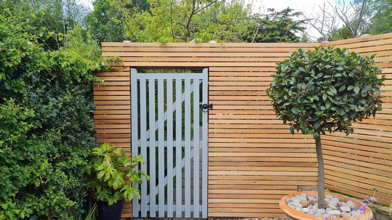 Wooden horizontal slat fence with a gray central gate; dense hedge to the left and a rounded potted tree in a rock-filled pot to the right.