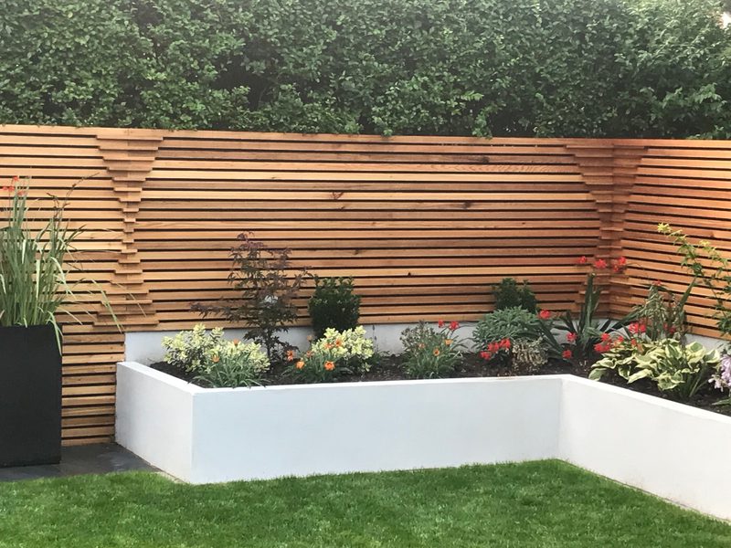 L-shaped white planter with colorful flowers before a horizontal wooden slat fence; a tall black planter on the left and a green lawn in front.