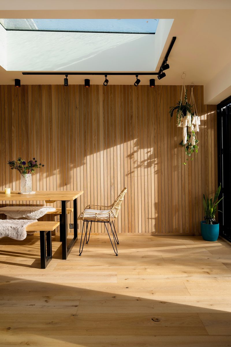 Sunlit dining nook with a wood slat wall, skylight, track lighting, a wooden table with benches and a vase of flowers, a wire chair, and hanging plants.
