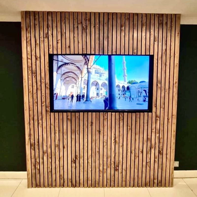 Flat-screen monitor inset in a vertical wood slat wall showing a sunlit courtyard with arches, columns, and a tall minaret.
