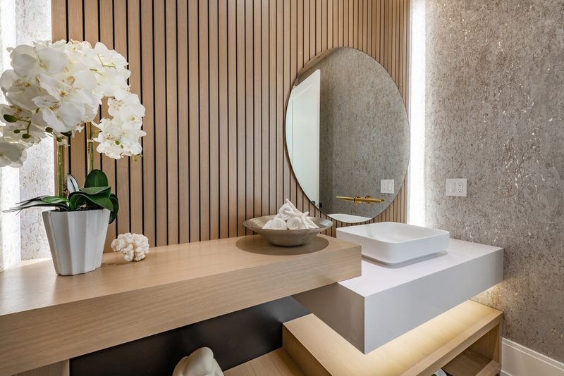 Modern bathroom vanity with round mirror, wooden slat wall, white vessel sink, gold faucet, orchid plant, and a bowl of towels.