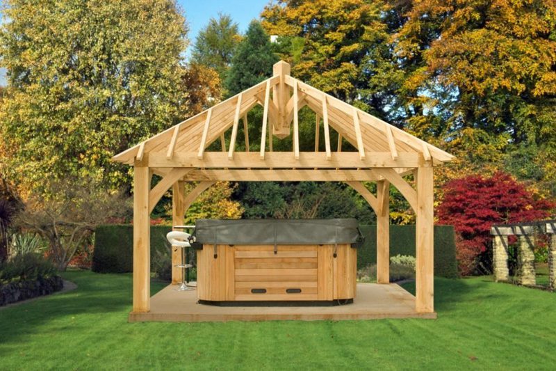 Wooden open-frame gazebo over a hot tub with a dark cover in a green lawn backyard, surrounded by autumn-colored trees.