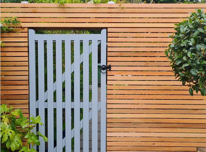 Light gray vertical-slat gate in a horizontal wooden fence, with a black latch. Behind and beside it are green plants, a small path, and a shrub on the right.