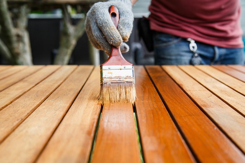 Gloved hand with a paintbrush applying stain to wooden deck planks outdoors; a person in a red shirt and jeans stands in the background.