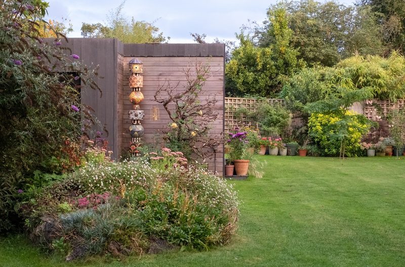 A tidy backyard garden with a wooden wall featuring stacked decorative birdhouses, a colorful flower bed in the foreground, and pots along the fence.