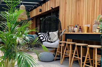 Cozy outdoor lounge with a wooden slat wall, hanging wicker chair, striped cushions, plants, string lights, and a bar with stools and bottles.