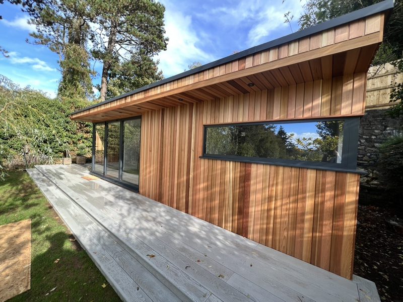 Modern wooden cabin with vertical planks, a large glass sliding door, a long side window, and a wide deck under a sloped roof, surrounded by trees.