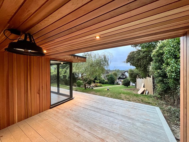 Wood-paneled covered patio with a sliding glass door opening onto a wooden deck and a grassy backyard with trees and a fence.