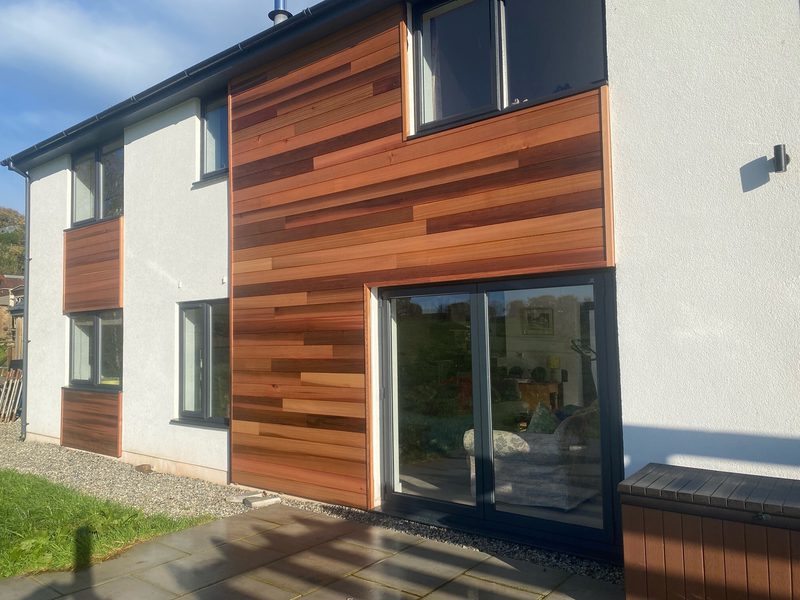 Modern white house with horizontal wooden cladding in warm brown tones, large sliding glass door, and multiple windows; gravel path and lawn in front.
