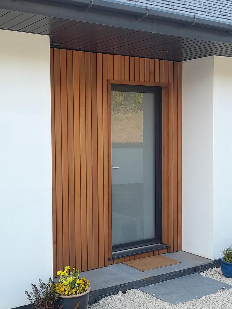 Entrance with a glass door framed by vertical wooden slats, white walls, and a dark overhang; brown mat, yellow flowers left, blue pot right.