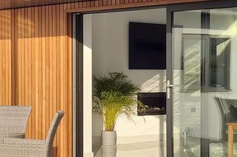 Sunny modern patio with a wicker round table and chairs, a wooden slat wall, a metal post, and large glass doors opening to an interior with a fireplace and plants.