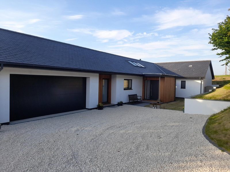 Modern, single-story home with a large black garage door, white walls, wooden entry panel, and a gravel driveway; bench outside and a blue sky above.