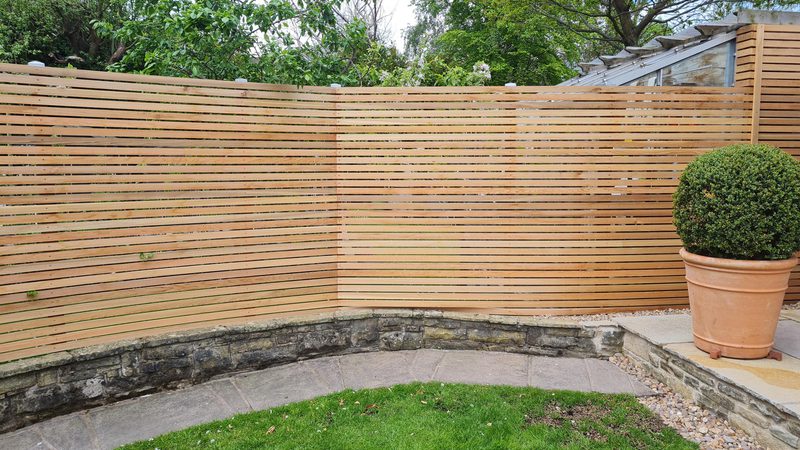 Curved horizontal wooden slat fence surrounding a small garden with a stone retaining wall, a round topiary in a terracotta pot, and a paved patio with grass.
