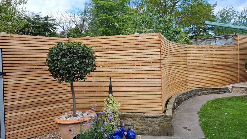 Curved horizontal wooden fence enclosing a garden with a potted topiary, purple flowers, stone edging, and a green lawn.