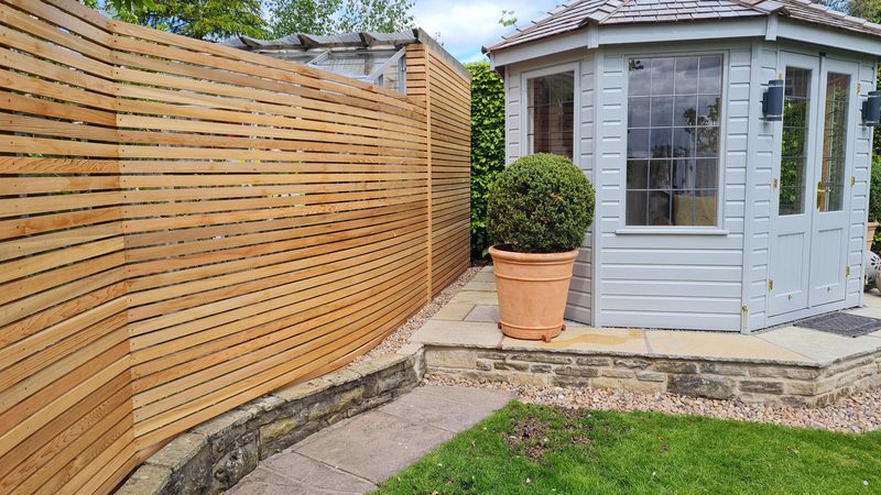 A backyard with a light gray garden shed with glass doors, a long horizontal wooden slatted fence on the left, a potted topiary, stone patio, and green lawn.