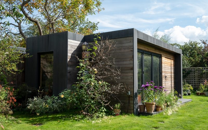A modern wooden garden shed with dark-framed sections and large glass doors, surrounded by potted plants on a green lawn under a blue sky.