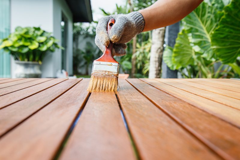 Close-up of a gloved hand painting a wooden deck with a brush; outdoor garden with green plants and a modern house in the background.