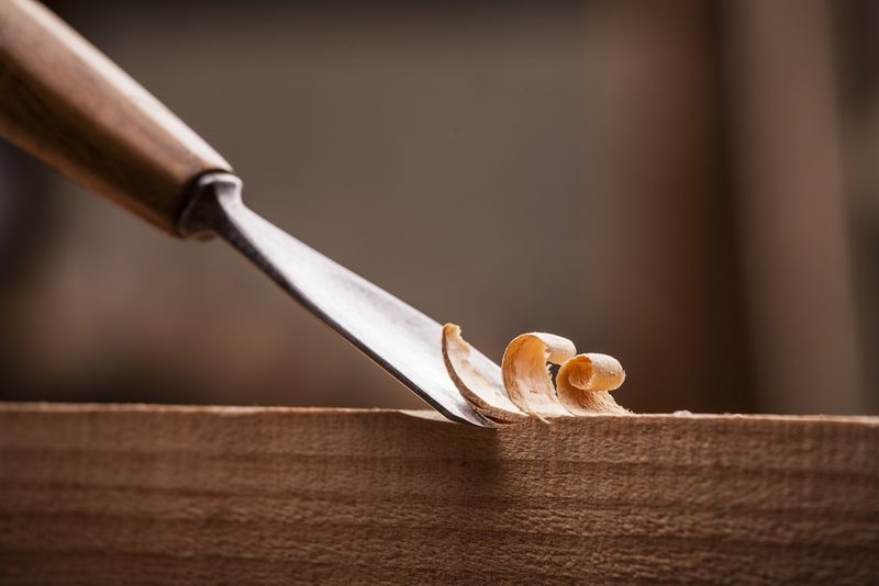 Close-up of a metal chisel slicing wood, with curling wood shavings resting on a wooden plank.