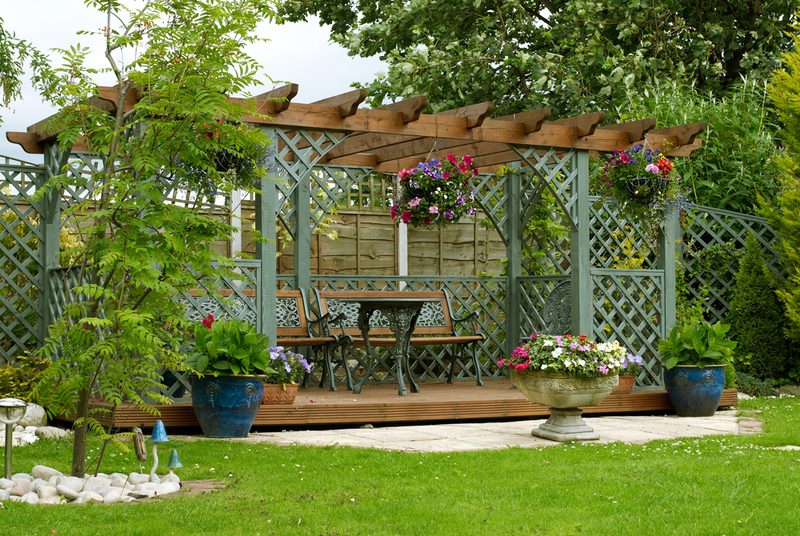 Garden pergola with lattice panels, a wooden bench and table, hanging baskets, and colorful potted flowers; lush lawn with trees and a stone planter.