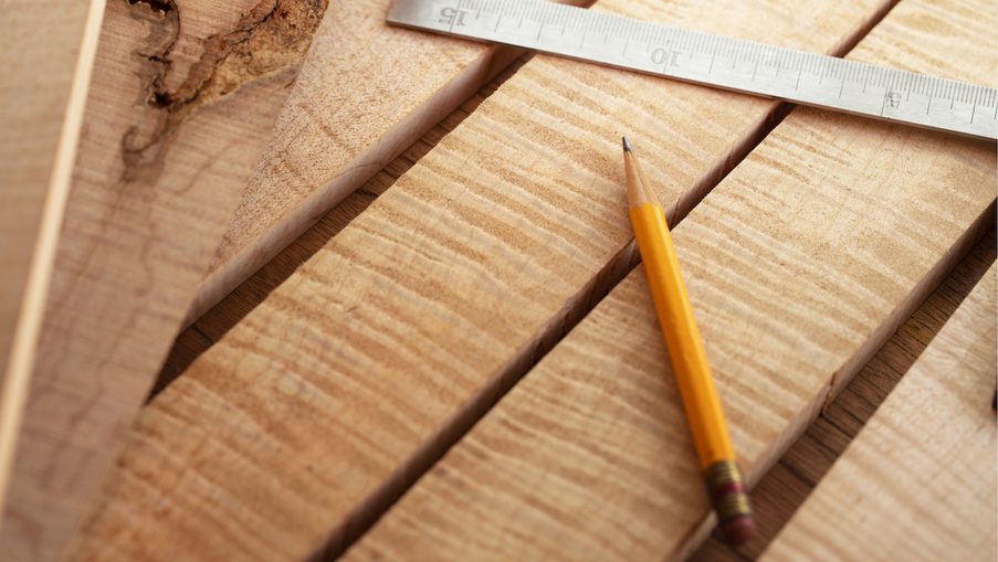 Close-up of light wood planks with a yellow pencil and a metal ruler laid diagonally across them.
