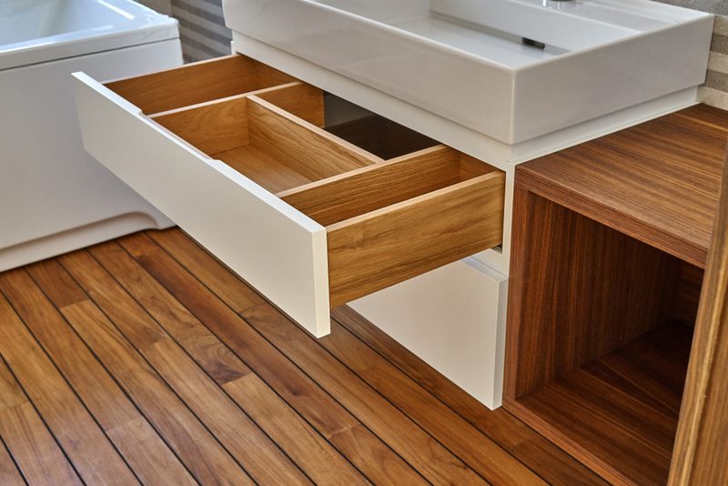 Partially pulled-out wooden drawer in a white modern bathroom vanity beneath a rectangular sink, with warm wood flooring and a nearby wooden cabinet.