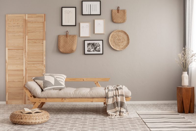 Neutral living room with a wooden daybed, cushions, plaid throw, woven pouf, screen divider, wall frames and baskets, and a vase of dried stems.