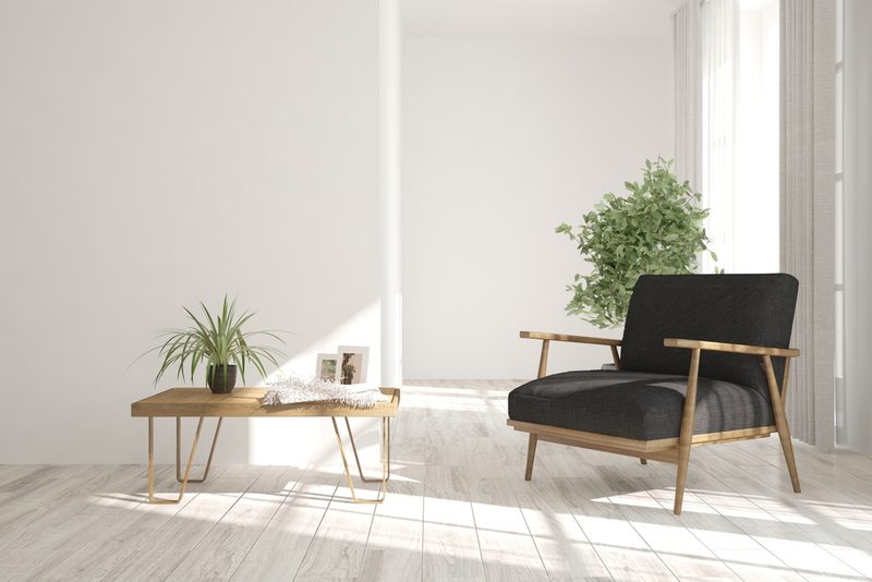Bright minimalist living room with a light wood coffee table, small plant and photos, and a black cushioned armchair by a sunlit window and tall plant.