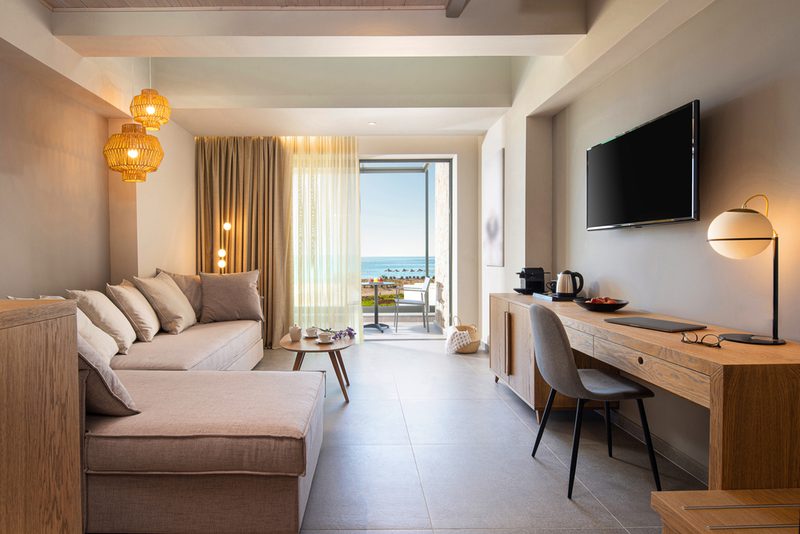 Bright modern hotel living area with a beige L-shaped sofa, wooden desk, wall TV, pendant lamps, and a balcony overlooking the ocean.