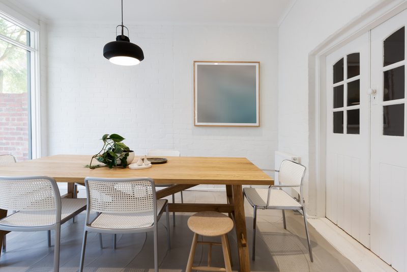 Bright minimalist dining room with a wooden table, white perforated chairs, a potted plant centerpiece, and a black pendant light.