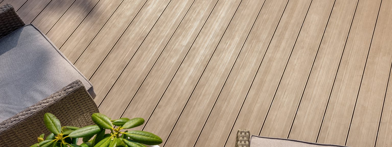 A wooden deck with long light-brown planks, a wicker chair with pale cushions in the lower left, and a potted green plant nearby.