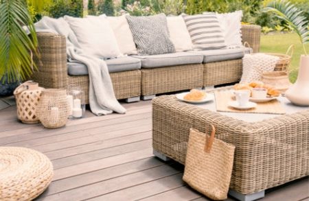 Sunny outdoor wicker sofa on a wooden deck with beige cushions, a woven coffee table, baskets, and plates of tea and pastries.