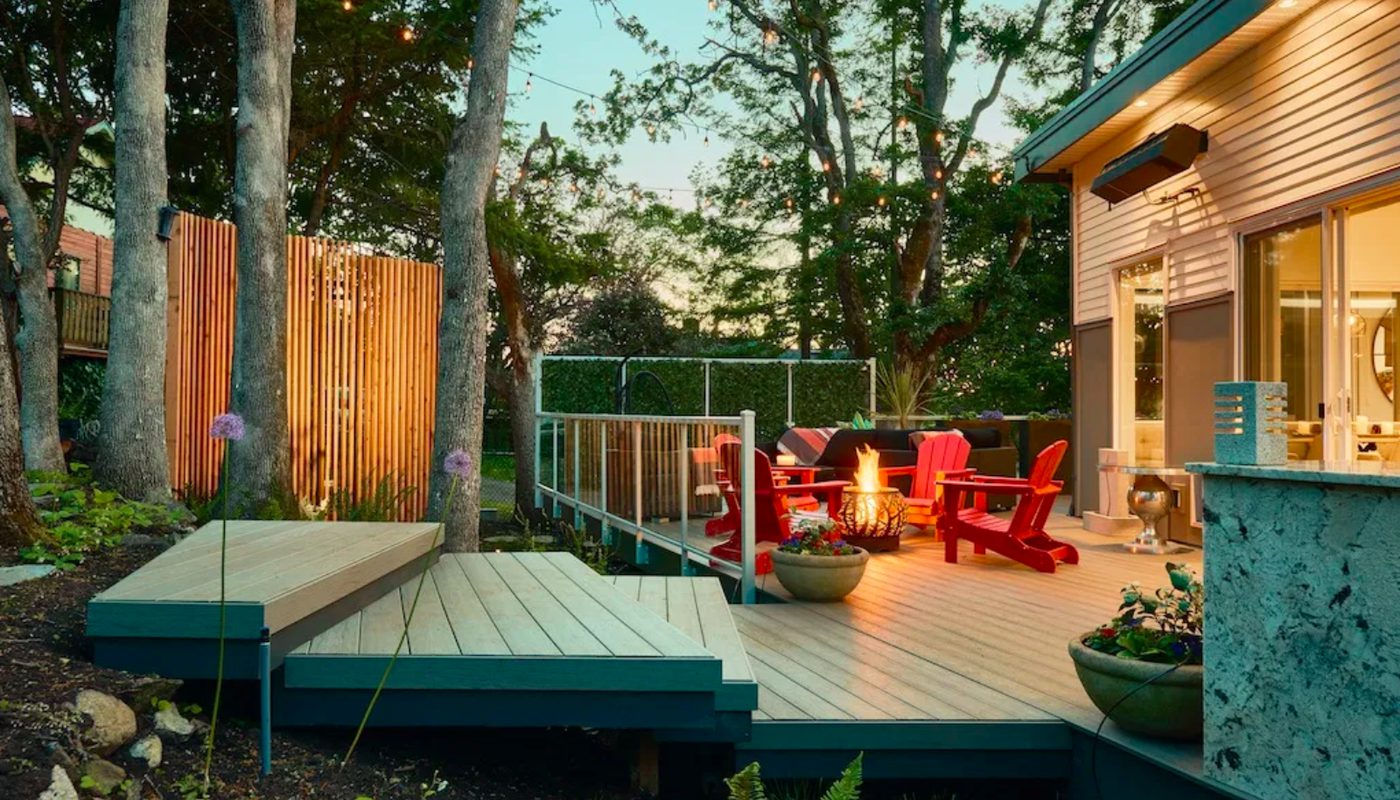 Backyard deck at dusk with string lights, trees, a glowing fire pit, and red Adirondack chairs around the seating area by the house patio.