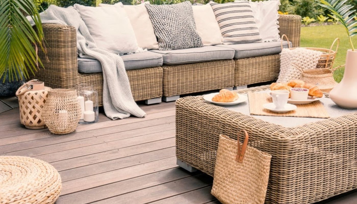 Outdoor wicker sofa with cushions on a wooden deck, a matching wicker coffee table with croissants and cups, baskets, candles, and greenery.