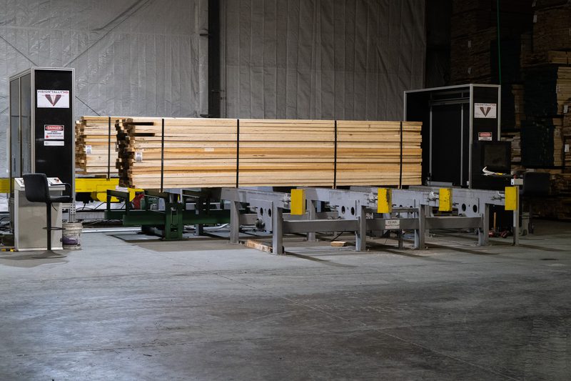 Industrial lumber facility with stacks of wooden boards on a metal conveyor, yellow safety blocks at the ends, a control station on the left, and tall storage in the back.