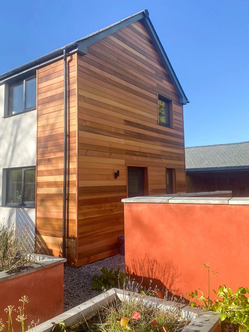 Two-story house with warm wood siding on the right and a white facade on the left, a red-orange privacy wall in the foreground, blue sky above.