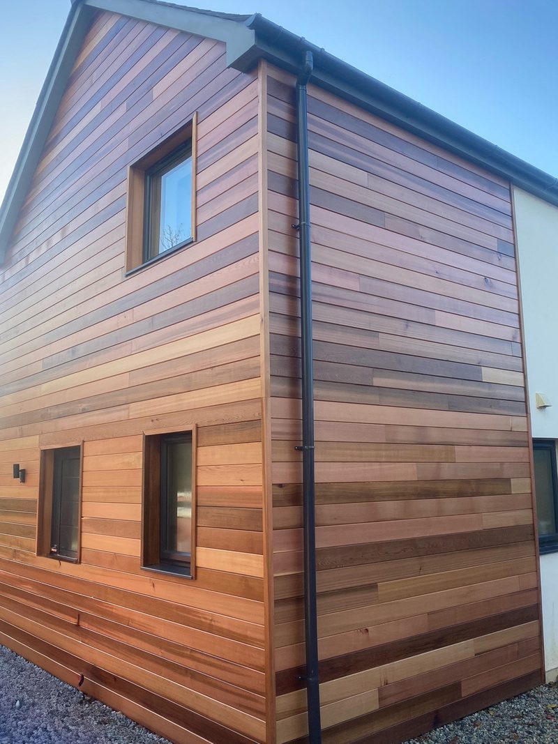 Two-story house with wooden siding in natural tones, two windows on the lower floor and one on the upper, black gutter and downspout, blue sky.