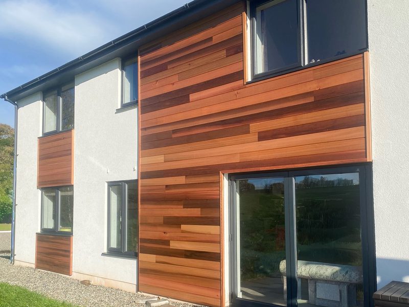 Modern two-story house with white exterior and horizontal brown wooden cladding, large glass door and windows, gravel path and blue sky.