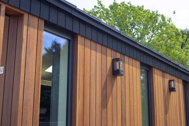 Exterior of a modern building with vertical wooden panels, large glass doors, two black wall lanterns, and a green tree in the background.