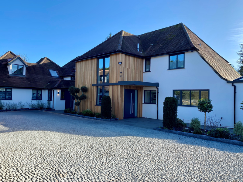 Modern white residence with a wooden central facade, blue entry door, large window, and sculpted evergreen bushes; pebble driveway under a blue sky.