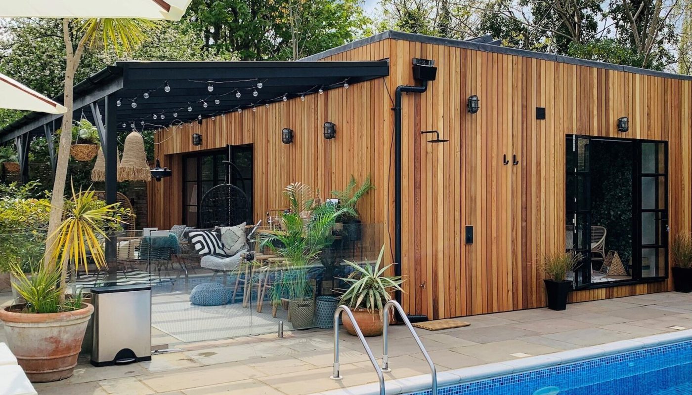 Wood-clad poolside deck with glass doors and string lights, cozy seating, and potted plants around a blue-tiled pool.
